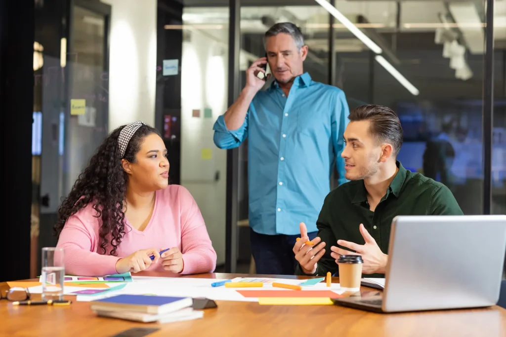 A team of three people in an office communicating. The manager is seen in the background on the phone.